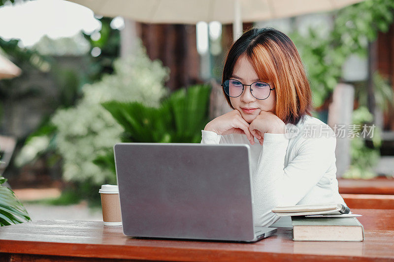 一个女人坐在桌子旁，手里拿着笔记本电脑和一杯咖啡。她戴着眼镜，脸上带着严肃的表情。这个场景表明她正在工作或学习