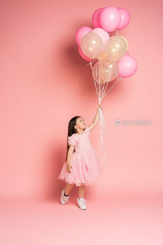 快乐的庆祝生日派对与迷人的可爱的小女孩在薄纱衣服微笑着对着相机孤立在粉红色的背景气球飞行庆祝。迷人的微笑，表达幸福