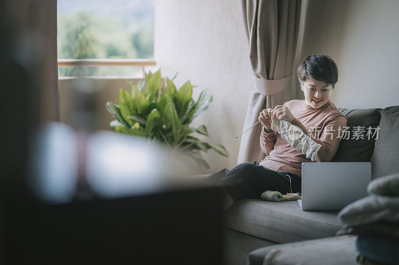 一个亚洲华人中年妇女在客厅编织在她的空闲时间，休闲活动和学习从她的笔记本电脑上网