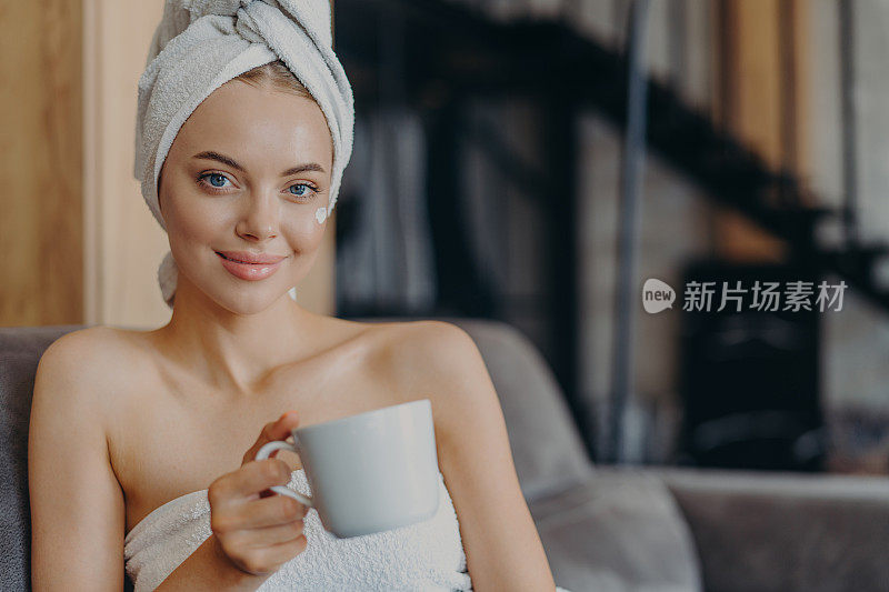 可爱的女人有着自然的妆容和健康的皮肤，露着肩膀，裹着浴巾，喝着茶或咖啡，坐在舒适的沙发上靠在室内。人,卫生,放松