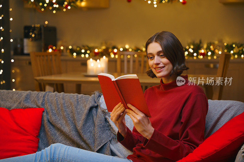 圣诞节前，一个女孩坐在带着圣诞彩灯的沙发上，微笑着读着一本书
