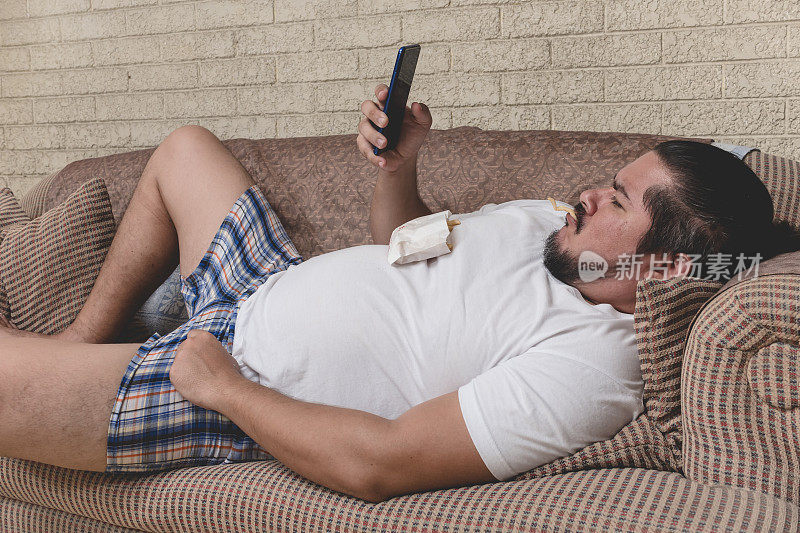 一个又懒又胖的流浪汉一边吃薯条一边看视频或在手机上浏览社交媒体。沙发上的土豆。