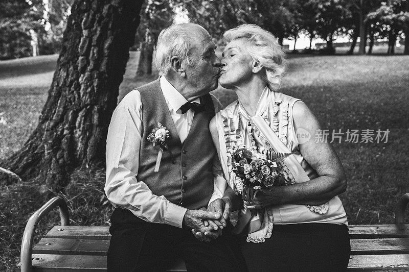 一对老夫妇坐在绿色公园的长凳上。祖母和祖父在金婚纪念日庆典上。五十年在一起。浪漫的老人是亲吻和拥抱。黑色和白色。