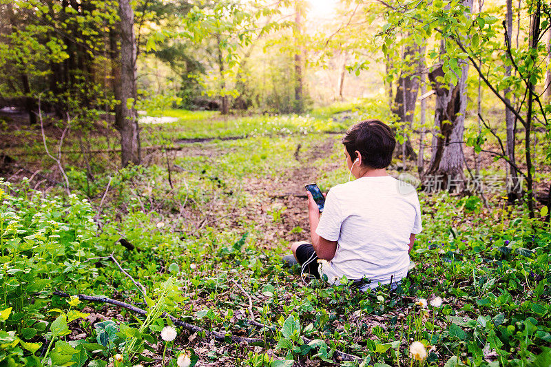一个十来岁的小男孩坐在森林里用耳机打电话。