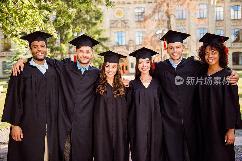 六个成功的快乐的多民族迷人的年轻毕业生，穿着黑色长袍和帽子完成了他们的教育，微笑和团结，背后是学院大楼，美好的阳光明媚的夏日