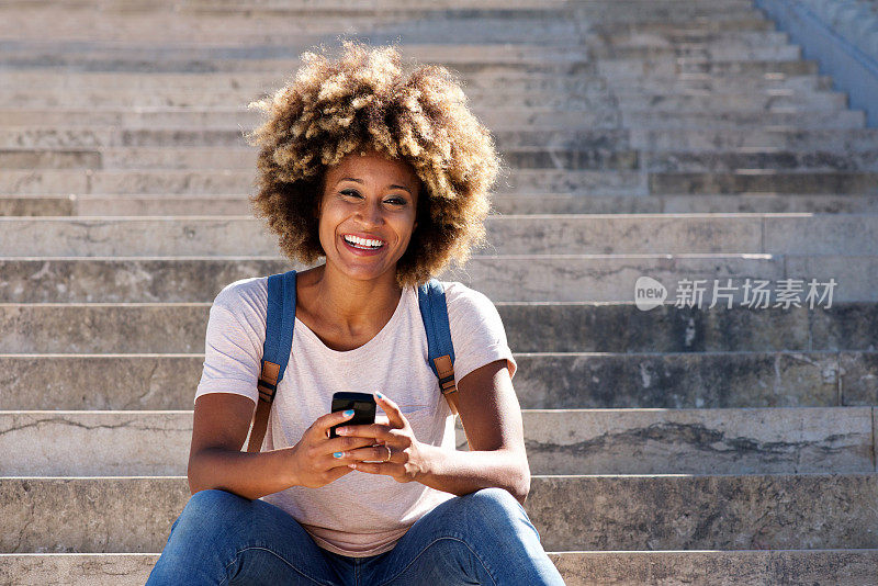 一名黑人妇女坐在台阶上拿着智能手机，在户外大笑