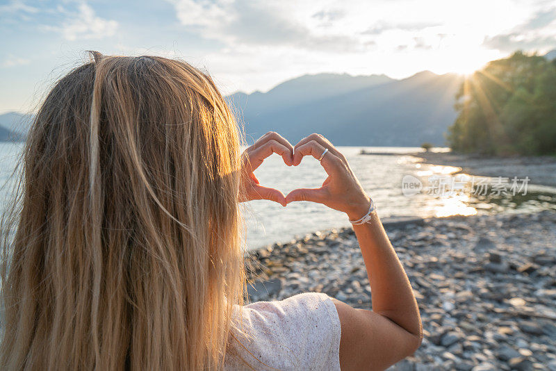 年轻的女子在美丽的山湖风景在夕阳下做一个心形的手指框架