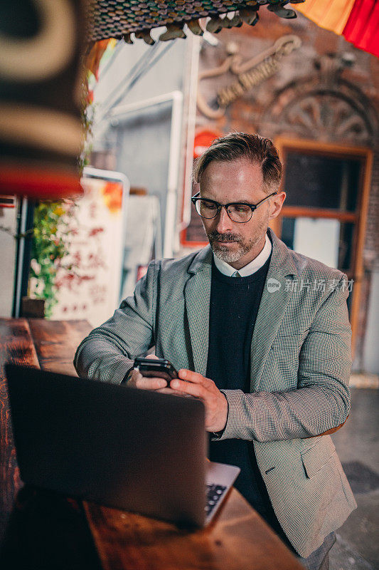 一个成熟的商人在路边的咖啡馆里使用手机和笔记本电脑