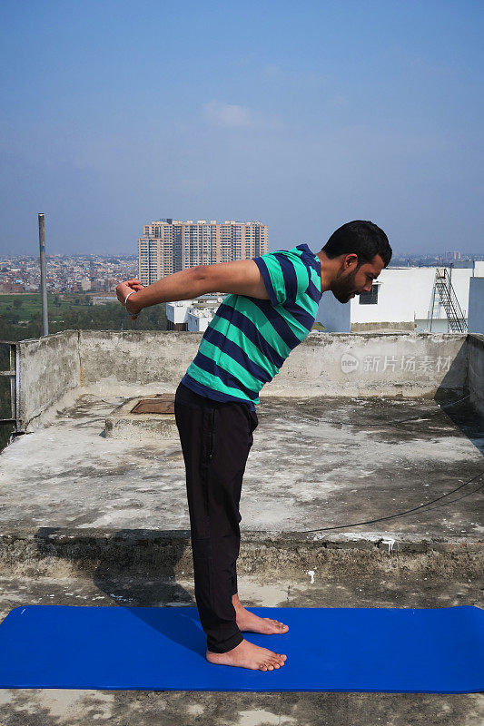 印度男子在屋顶上练习手握向前弯曲(Uttanasana)瑜伽姿势，德里，印度