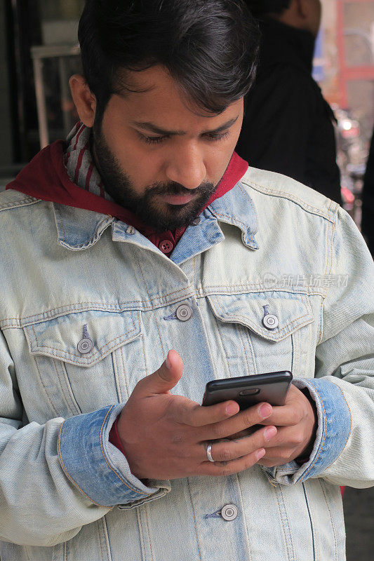 在印度德里，英俊的印度男子用手机看屏幕发短信，查看社交媒体和互联网，年轻的印度男子用手机交流，城市背景，连帽衫青年牛仔夹克