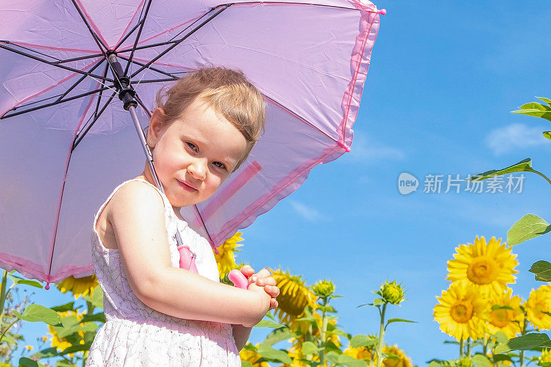 4岁的白人女孩撑着一把粉红色的雨伞挡住了阳光，一个孩子拿着向日葵花，微笑着看着镜头。