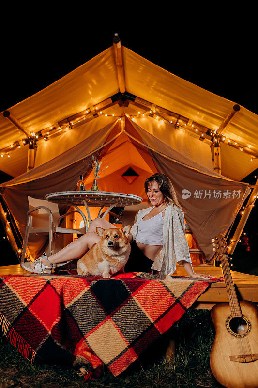 快乐的年轻女子与她的威尔士柯基彭布罗克狗放松在夏季晚上豪华露营附近舒适的篝火。户外休闲休闲的豪华露营帐篷。生活方式的概念