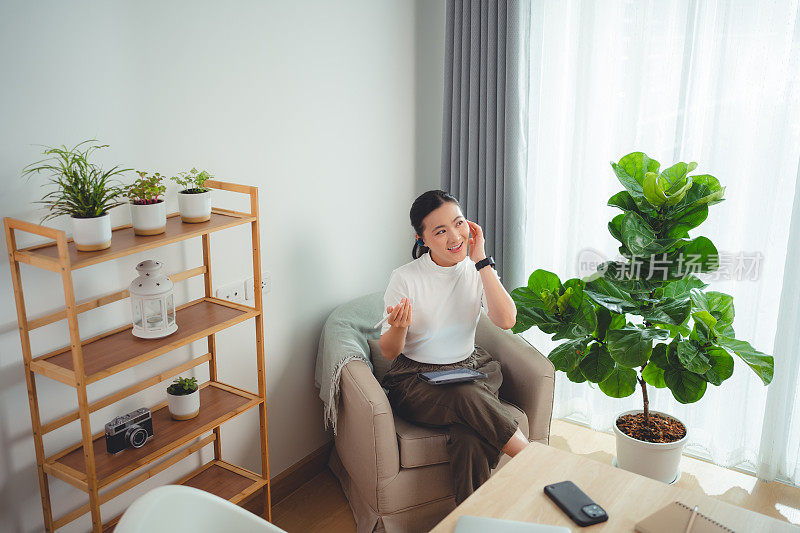 亚洲女性喜欢戴着耳机上网工作，在平板电脑上阅读数据，坐在家里的扶手椅上打电话。