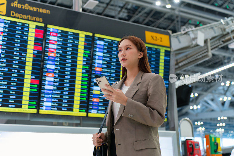 一名年轻的亚洲女子手持手提包和智能手机，站在机场候机楼的航班信息显示屏前。出差的商务人士。手持智能手机的年轻亚洲女性旅客在机场候机楼。