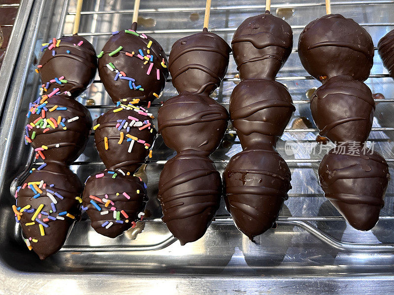 特写图片:一排排的木制串，每个串上有三个黑巧克力覆盖的草莓，上面装饰着巧克力和糖屑，放在金属托盘架上，玻璃镶板的面包店货架展示，高架视图