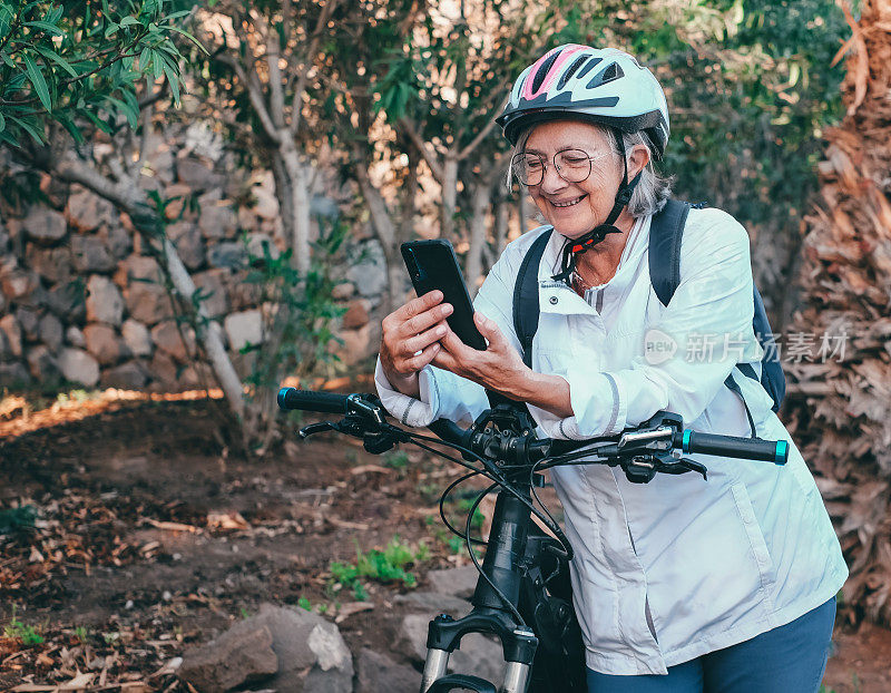 带着自行车头盔的微笑老年妇女在户外活动中骑着电动自行车使用手机。一位老妇人在公园里进行健康活动