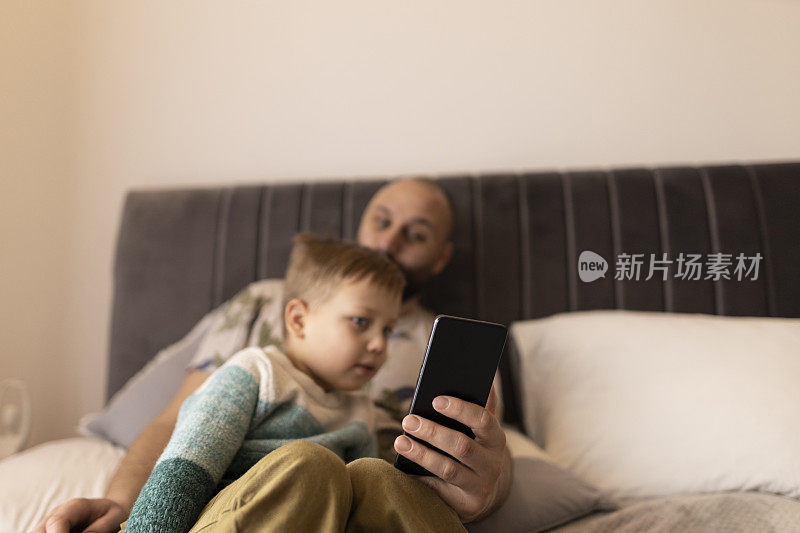 爸爸和小儿子笑着看着手机上的照片。他们坐在卧室的床上拍照。父亲拥抱儿子。他们做鬼脸。一个黑发浓密的男人和一个金发碧眼的男孩。焊接时间