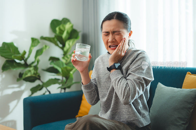 亚洲女性坐在客厅的沙发上，喝了凉水，摸了摸脸颊，就会感到牙齿敏感。