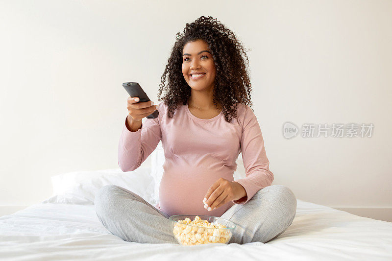 穿着粉色衬衫的黑人孕妇拿着遥控器享受着爆米花