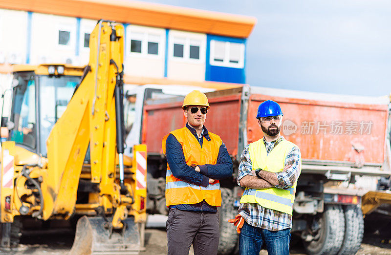 两名工程师在建筑工地的卡车和机器旁摆姿势