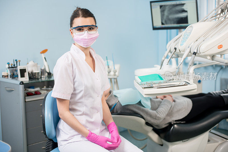 年轻女牙医与病人在牙科诊所的肖像。医生戴眼镜，口罩，白色制服和粉红色手套。背景屏幕上显示的是病人的牙齿。牙科