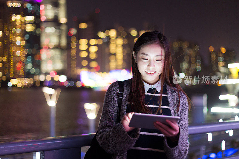 年轻美丽的亚洲女人晚上在现代城市玩数字平板电脑