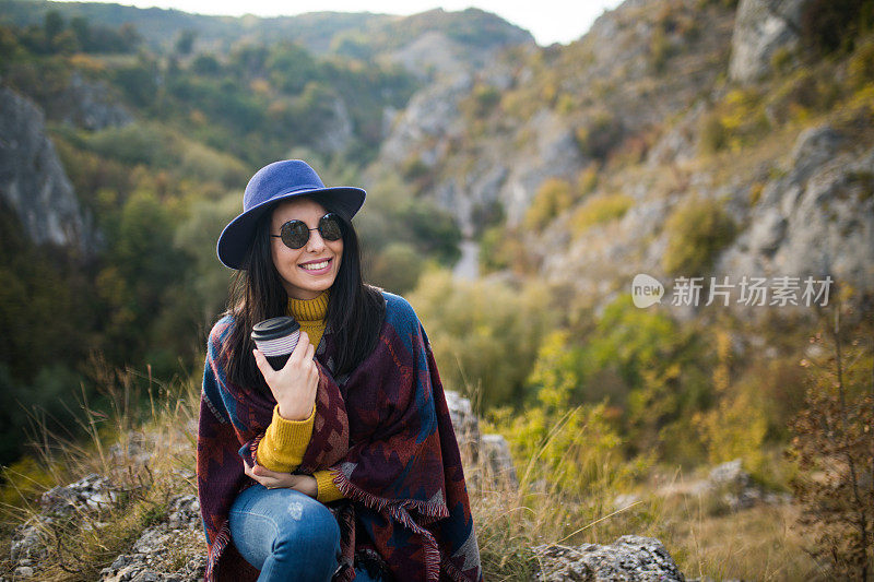 女旅行者一边喝着咖啡，一边欣赏着山景。一个年轻的女游客喝着一杯热饮，欣赏着山上的风景。徒步旅行的概念