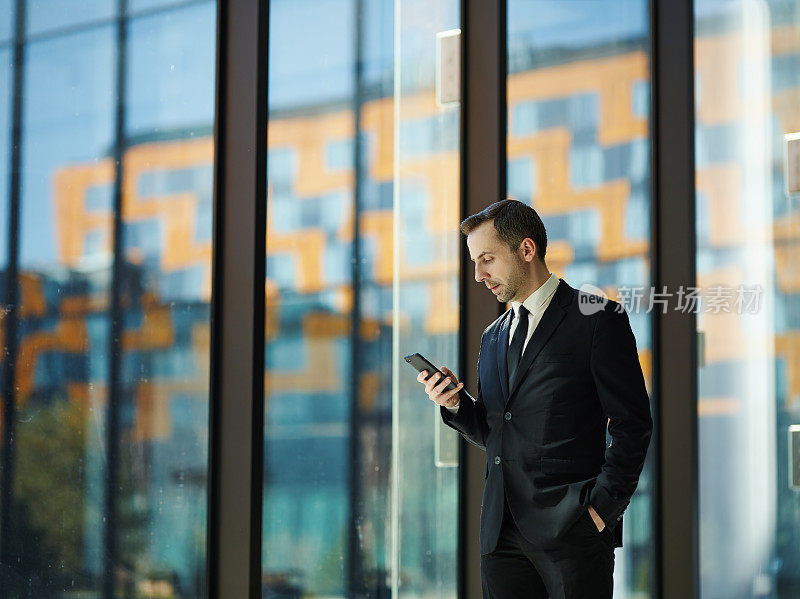 四分之三长度的肖像自信的经理在正装站在窗口与手在口袋和浏览手机，拷贝空间向左