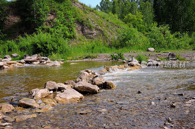 在夏季森林的山坡上有岩石海岸的一条浅河的河岸。