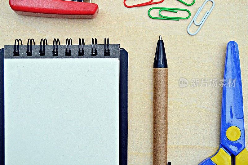 一个空白的白纸螺旋装订的记事本，连同一支钢笔，彩色回形针和塑料剪刀在木制背景
