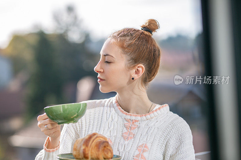 年轻女子坐在窗前吃牛角面包，喝咖啡休息