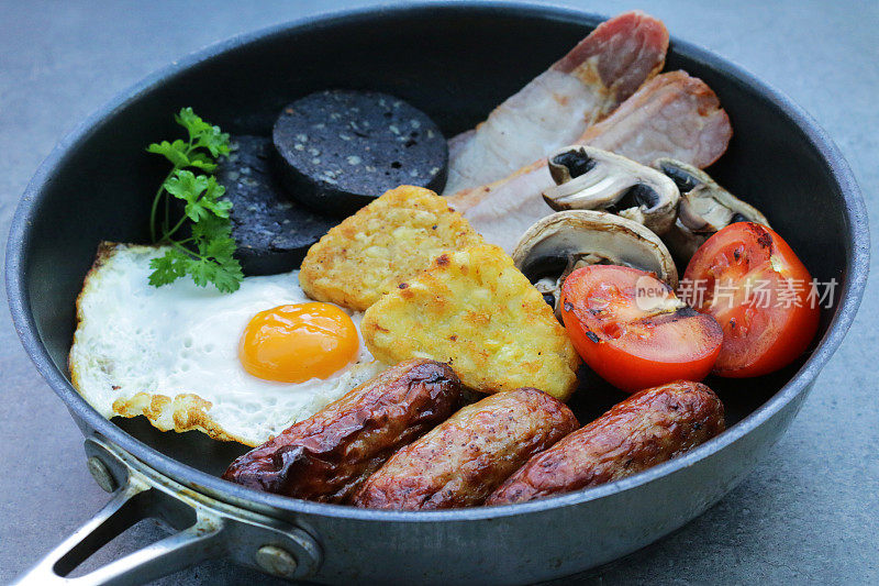 不粘锅的图像包含了完整的英国早餐的元素，煮猪肉香肠，土豆煎饼，西红柿，蘑菇片，培根片，煎蛋和黑布丁(血肠)