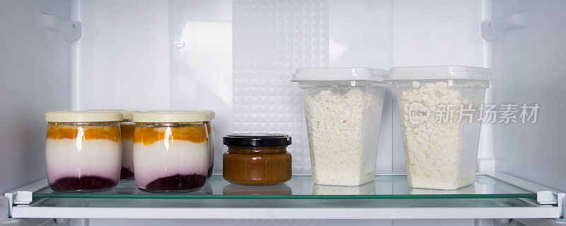 在冰箱的玻璃架子上，有罐装酸奶、水果和塑料盒里的碎软干酪