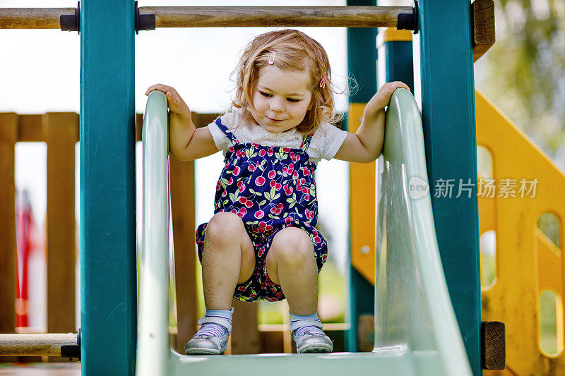 可爱的蹒跚学步的女孩在户外操场上玩滑梯。美丽的宝宝穿着五颜六色的短裤裤子在阳光温暖的夏天玩。孩子滑下来