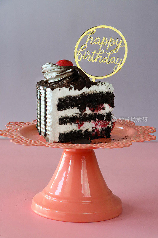 黑色森林蛋糕片的图像，三层蛋糕层被橙色蛋糕架上的鲜奶油隔开，生日快乐标志，管状鲜奶油花环，莫雷罗樱桃，豪华巧克力蛋糕，巧克力刨花，粉紫色背景，重点在前景