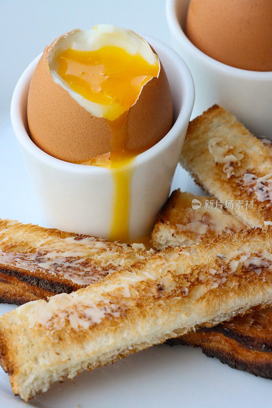 特写图像黄油吐司士兵与两个软煮鸡蛋在蛋杯在白盘子，顶部软煮鸡蛋与溏心黄色蛋黄，重点在前景