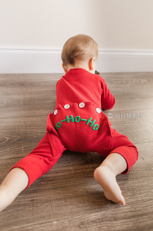 一个11个月大的圣诞男婴长着12只脚趾，穿着红色复古风格的圣诞睡衣，睡衣底部写着“HO-HO-HO”，在木地板上玩耍，为他的生日和2022年的假期做准备