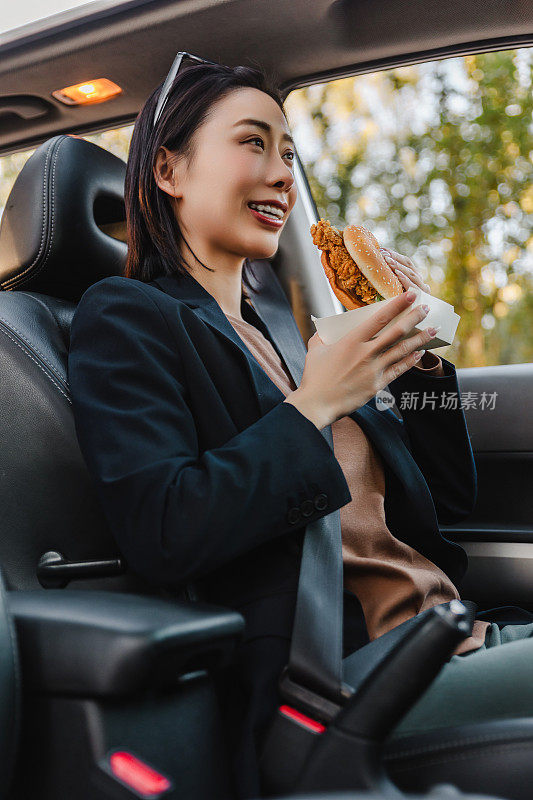亚洲女人在车里休息，吃汉堡。一个年轻成功快乐的女人正在开车。