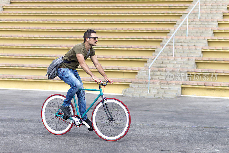 侧视图，年轻男子戴着太阳镜，背着肩包，骑着自行车。