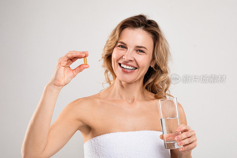 美容维生素。快乐的中年女士拿着一杯水和药丸
