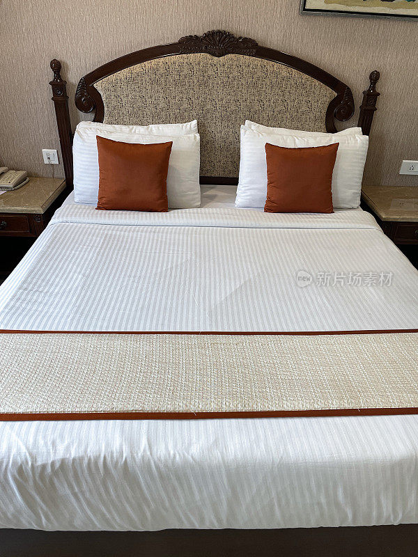 一叠白色酒店枕头倚在织物床头板上，白色羽绒被床罩，青铜色靠垫，床脚处的床轮，聚焦于前景