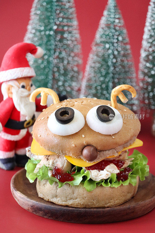 圣诞驯鹿设计的圣诞老人形象火鸡和蔓越莓三明治，芝麻籽汉堡面包上面有洋葱和去核的黑橄榄“眼睛”，盐椒盐脆饼“鹿角”和巧克力球“鼻子”，塑料冷杉树，红色背景