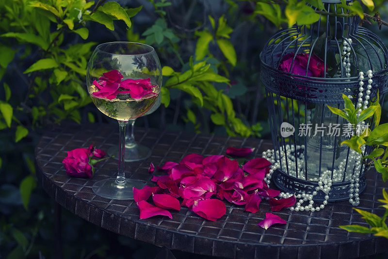 花园的桌子上有一杯酒和红玫瑰花瓣。鸟笼和珍珠珠。复古时尚的黑暗照片。