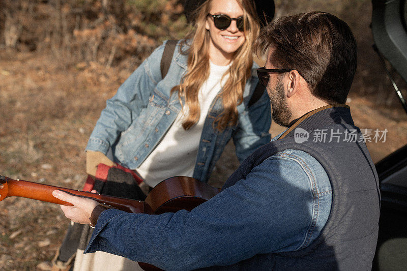 在他们的公路旅行冒险中，一个男人在汽车后备箱旁给他的未婚妻弹吉他