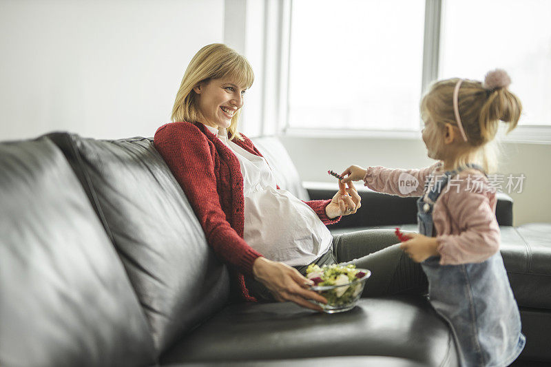 怀孕的母亲和女儿吃着健康的沙拉，在家里的客厅里玩耍