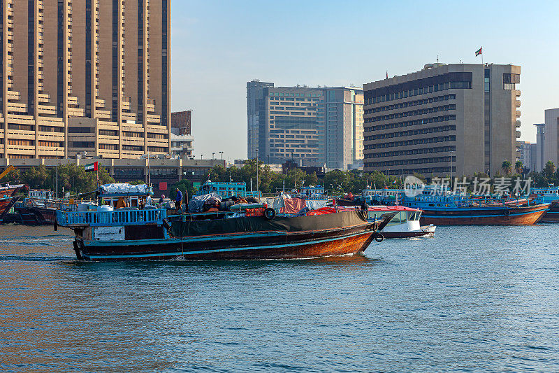 阿拉伯联合酋长国迪拜——晨曦中，一艘满载的传统阿拉伯单桅帆船驶入迪拜河——背景中，在德拉一侧，单桅帆船彼此并排停泊。在遥远的背景，办公室和住宅建筑。