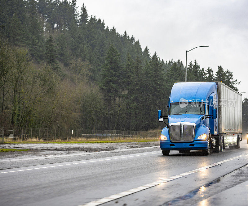 专业的蓝色大卡车半挂车运送商业货物在干式货车半挂车运行在晚上潮湿的雨天道路与森林的一边