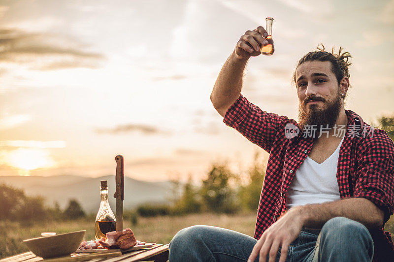 自然的守护者。一个在山上的男人，一个伐木工人，喝着自制的白兰地，吃着自制的食物，而在他身后是美丽的日落，休息辛苦的工作。