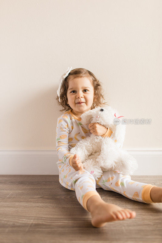 2019冠状病毒病期间，两岁蹒跚学步的小女孩穿着睡衣和蝴蝶结在家拥抱和亲吻她毛茸茸的白色狗玩具