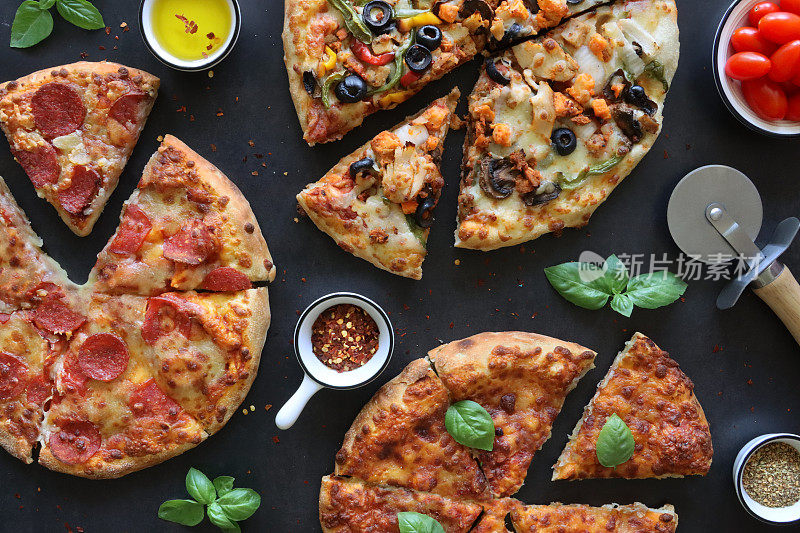 新鲜出炉的玛格丽塔披萨和意大利辣香肠披萨，金色磨碎的马苏里拉奶酪，浓郁的番茄沙司和意大利辣香肠片，蔬菜披萨(黄色和绿色的柿子椒和黑橄榄)，披萨轮，黑色背景，高架视图